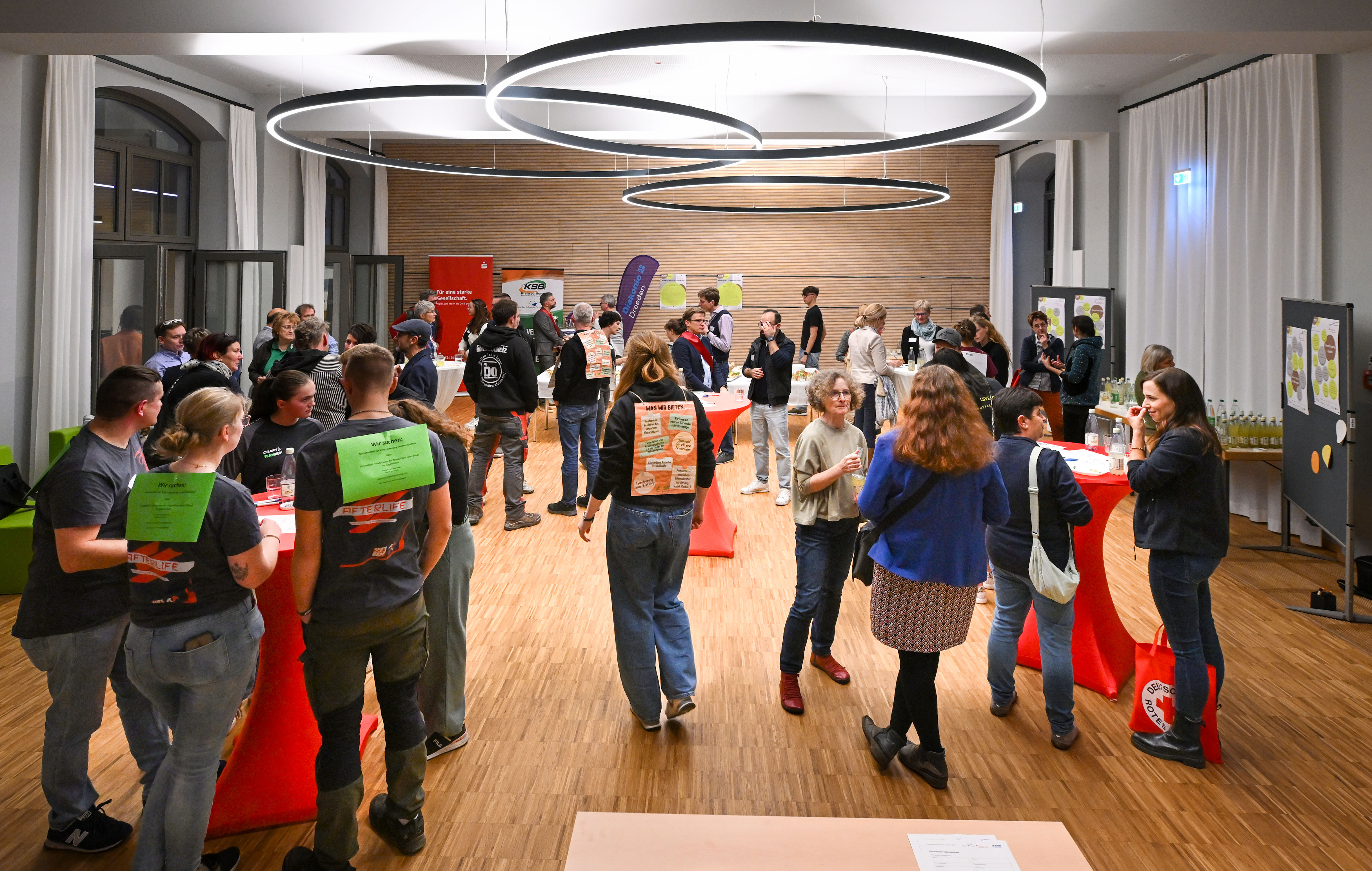 KI generiert: Das Bild zeigt eine belebte Indoor-Veranstaltung mit vielen Menschen, die sich in Gruppen unterhalten und an Stehtischen mit roten Tischdecken stehen. Im Raum gibt es mehrere Informationsstände und eine moderne Beleuchtung.