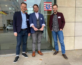 KI generiert: Das Bild zeigt eine Podiumsdiskussion mit vier Personen, die auf der Bühne sitzen und sich unterhalten, während eine Präsentation oder ein Video auf einer Leinwand im Hintergrund läuft. Der Hauptinhalt des Bildes ist die Diskussion oder das Panel auf der Bühne.
