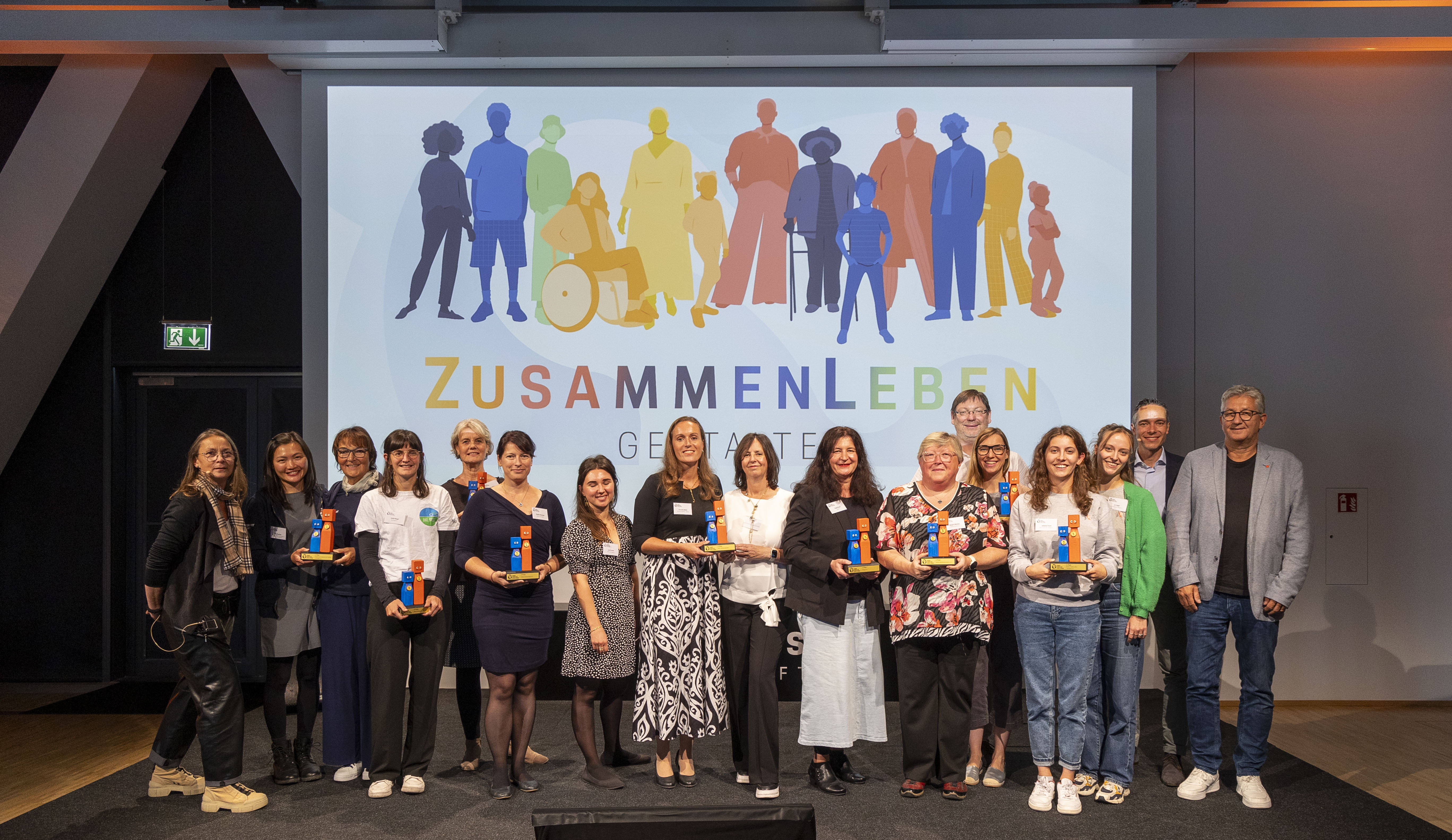 KI generiert: Das Bild zeigt eine Gruppe von Personen, die auf einer Bühne stehen und gemeinsam in die Kamera lächeln. Im Hintergrund ist ein Banner mit dem Schriftzug "ZUSAMMENLEBEN GESTALTEN" und einer Illustration von verschiedenen Menschen zu sehen.