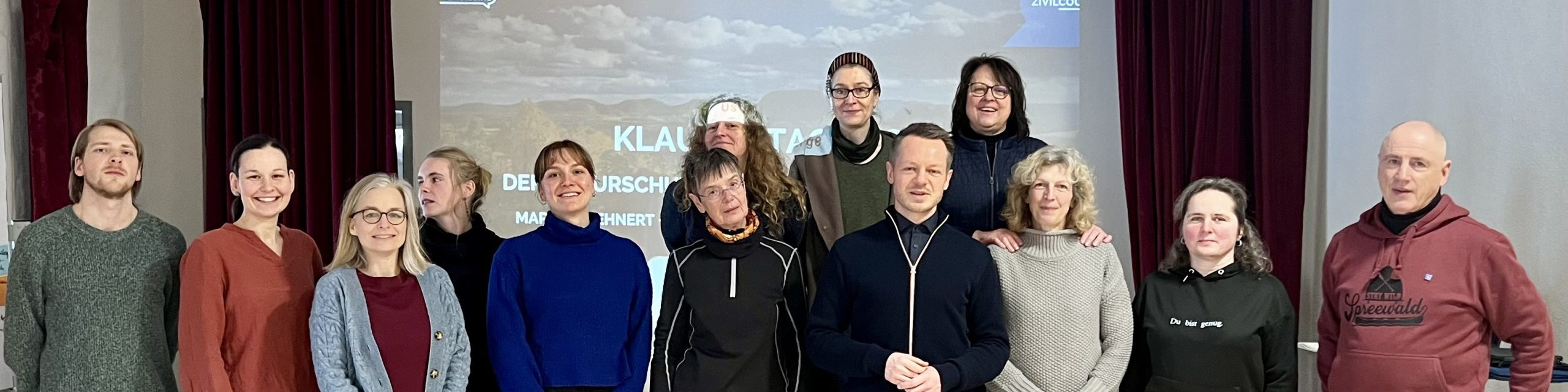 KI generiert: Das Bild zeigt eine Gruppe von Personen, die vor einem Projektorbildschirm mit der Aufschrift "KLAUSURTAG" posieren. Der Hauptinhalt des Bildes ist eine Gruppenaufnahme, die vermutlich im Rahmen einer Klausurtagung oder eines ähnlichen Anlasses entstanden ist.