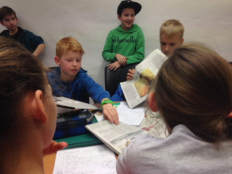 KI generiert: Das Bild zeigt eine Gruppe von Kindern, die gemeinsam an einem Tisch sitzen und Zeitungen lesen oder besprechen. Der Hauptinhalt des Bildes ist eine Lern- oder Gruppenaktivität, bei der die Kinder sich mit gedruckten Materialien beschäftigen.