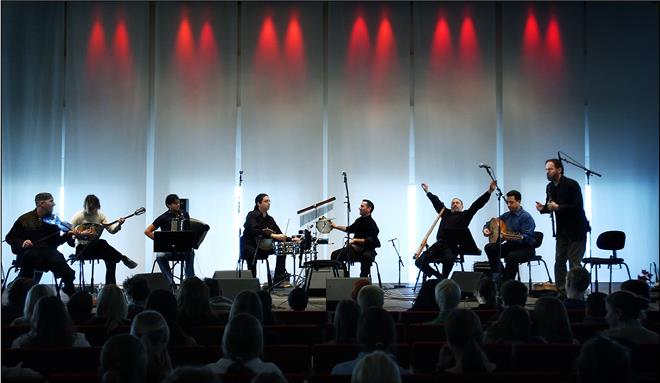KI generiert: Das Bild zeigt eine Gruppe von Musikern, die auf einer Bühne verschiedene Instrumente spielen, während ein Mann in der Mitte singt oder spricht. Im Vordergrund ist ein Publikum zu sehen, das der Darstellung aufmerksam folgt.