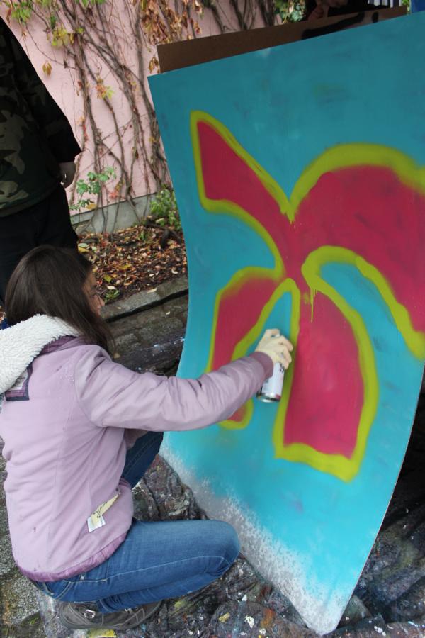 KI generiert: Das Bild zeigt eine Person, die auf einem großen, blauen Holzbrett ein Graffiti in Form einer roten Blume mit gelben Konturen sprüht. Die Szene spielt im Freien, möglicherweise in einem Park oder einer urbanen Umgebung.