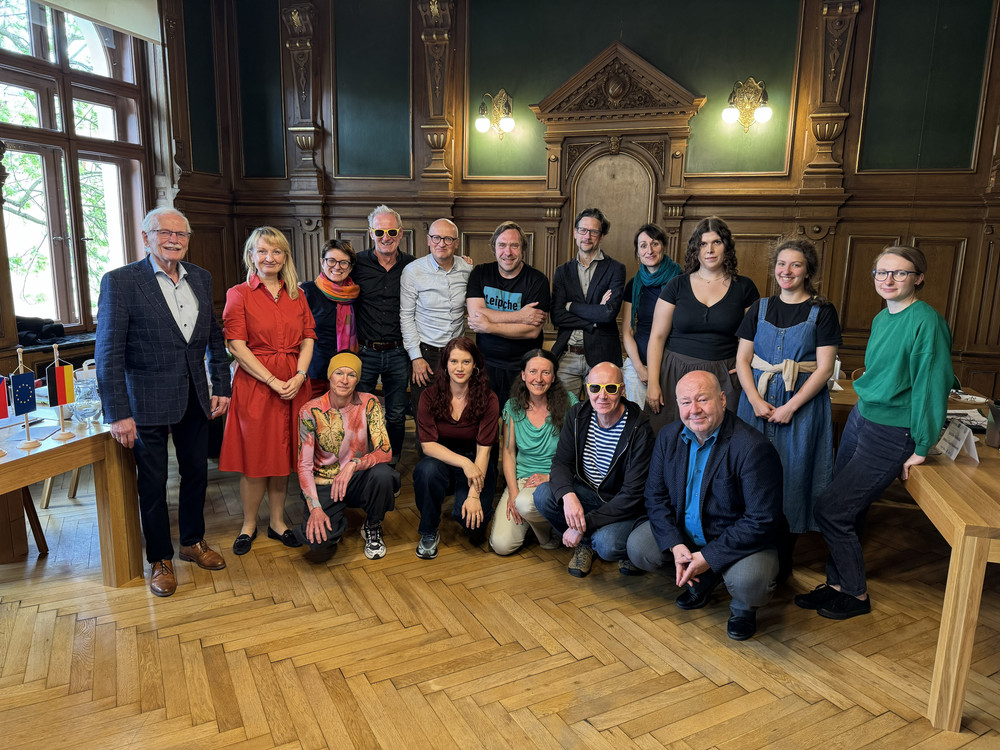 KI generiert: Das Bild zeigt eine Gruppe von Personen, die in einem Raum mit Holzvertäfelung und großen Fenstern posieren. Im Vordergrund sind zwei kleine Flaggen zu sehen, was darauf hindeuten könnte, dass es sich um eine offizielle oder internationale Zusammenkunft handelt.