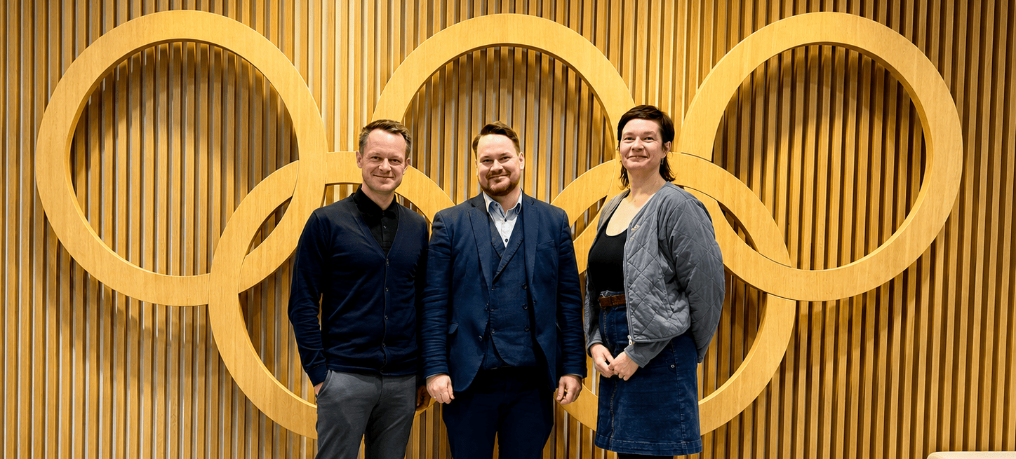 KI generiert: Das Bild zeigt drei Personen, die vor einer Holzvertäfelung mit den olympischen Ringen posieren. Der Hauptinhalt des Bildes ist die Darstellung der drei Individuen vor dem Symbol der Olympischen Spiele.