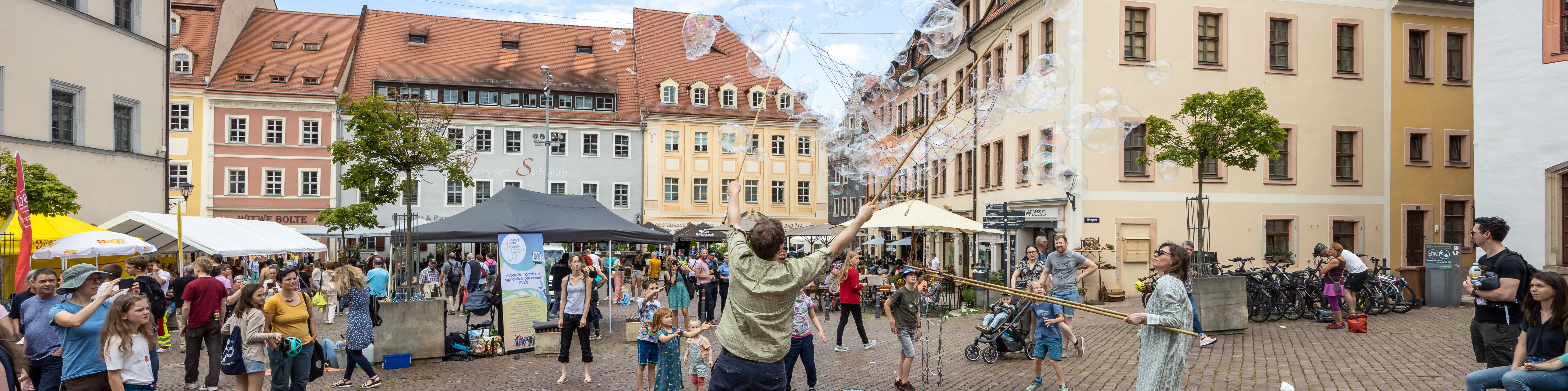 KI generiert: Das Bild zeigt eine lebendige Straßenveranstaltung in einer historischen Stadt mit farbenfrohen Gebäuden, bei der Menschen, darunter auch Kinder, Seifenblasen in die Luft pusten. Im Hintergrund sind Marktstände und eine fröhliche Menschenmenge zu sehen, die den Tag genießt.