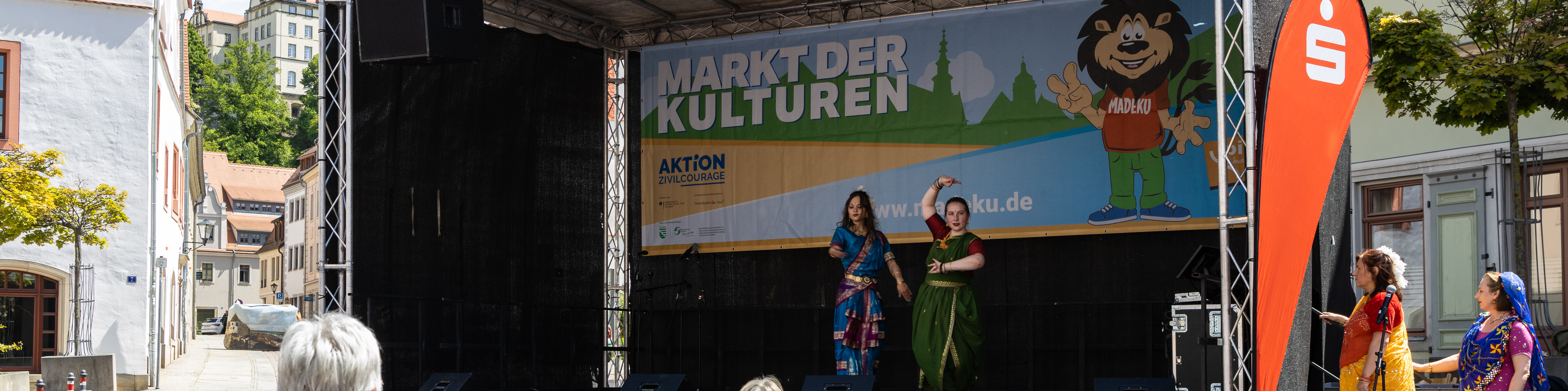 KI generiert: Das Bild zeigt eine Open-Air-Bühne mit der Aufschrift "Markt der Kulturen" und einer Website-Adresse, auf der zwei Frauen in traditioneller Kleidung tanzen. Im Hintergrund sind Gebäude und einige Zuschauer zu sehen.