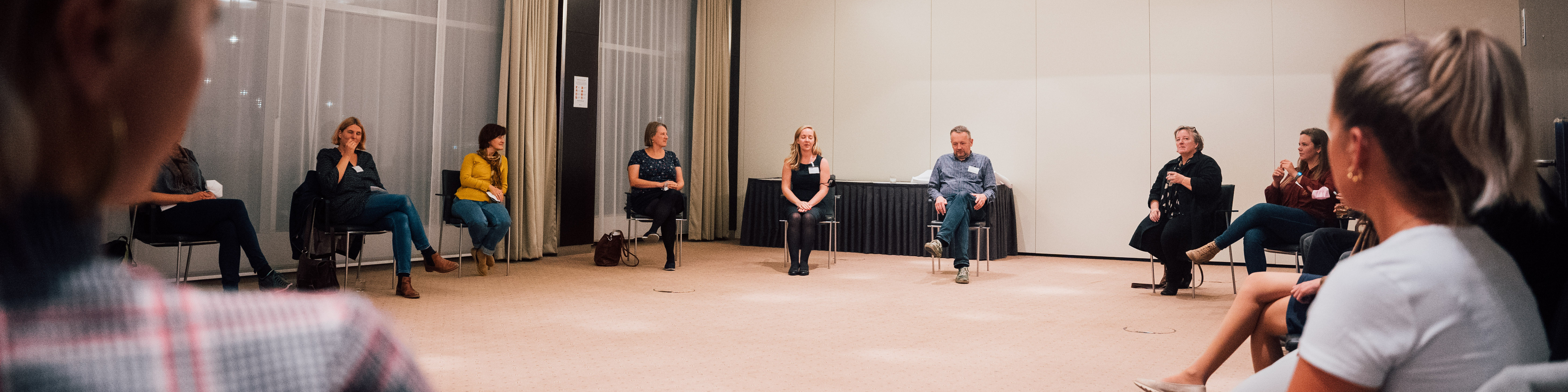 KI generiert: Das Bild zeigt eine Gruppe von Personen, die in einem hellen Raum auf Stühlen im Kreis sitzen und sich unterhalten. Der Hauptinhalt ist eine Diskussion oder ein Meeting in einer informellen Atmosphäre.