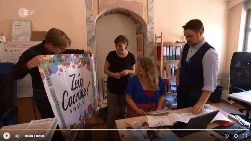 KI generiert: Das Bild zeigt vier Personen in einem Büro, die gemeinsam ein Plakat mit der Aufschrift "Zeig Courage! 19.09.2019" betrachten. Der Hauptinhalt des Bildes ist die Präsentation oder Besprechung des Plakats, möglicherweise im Rahmen einer Kampagne oder Veranstaltung.