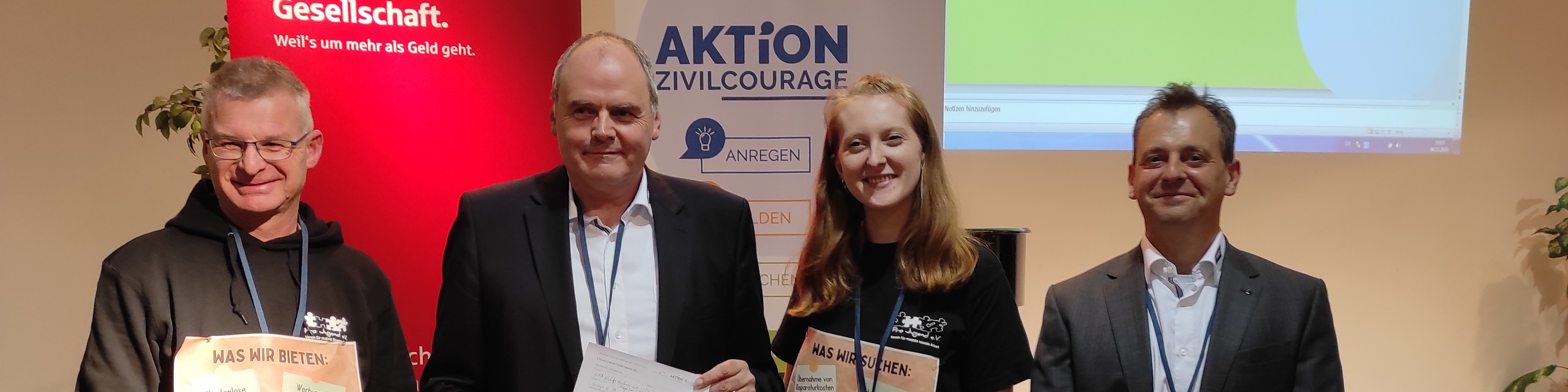 KI generiert: Das Bild zeigt vier Personen, die gemeinsam vor einem Banner mit der Aufschrift "AKTION ZIVILCOURAGE" stehen und Dokumente oder Preise in den Händen halten. Es scheint sich um eine Veranstaltung oder Auszeichnung im Zusammenhang mit Zivilcourage zu handeln.