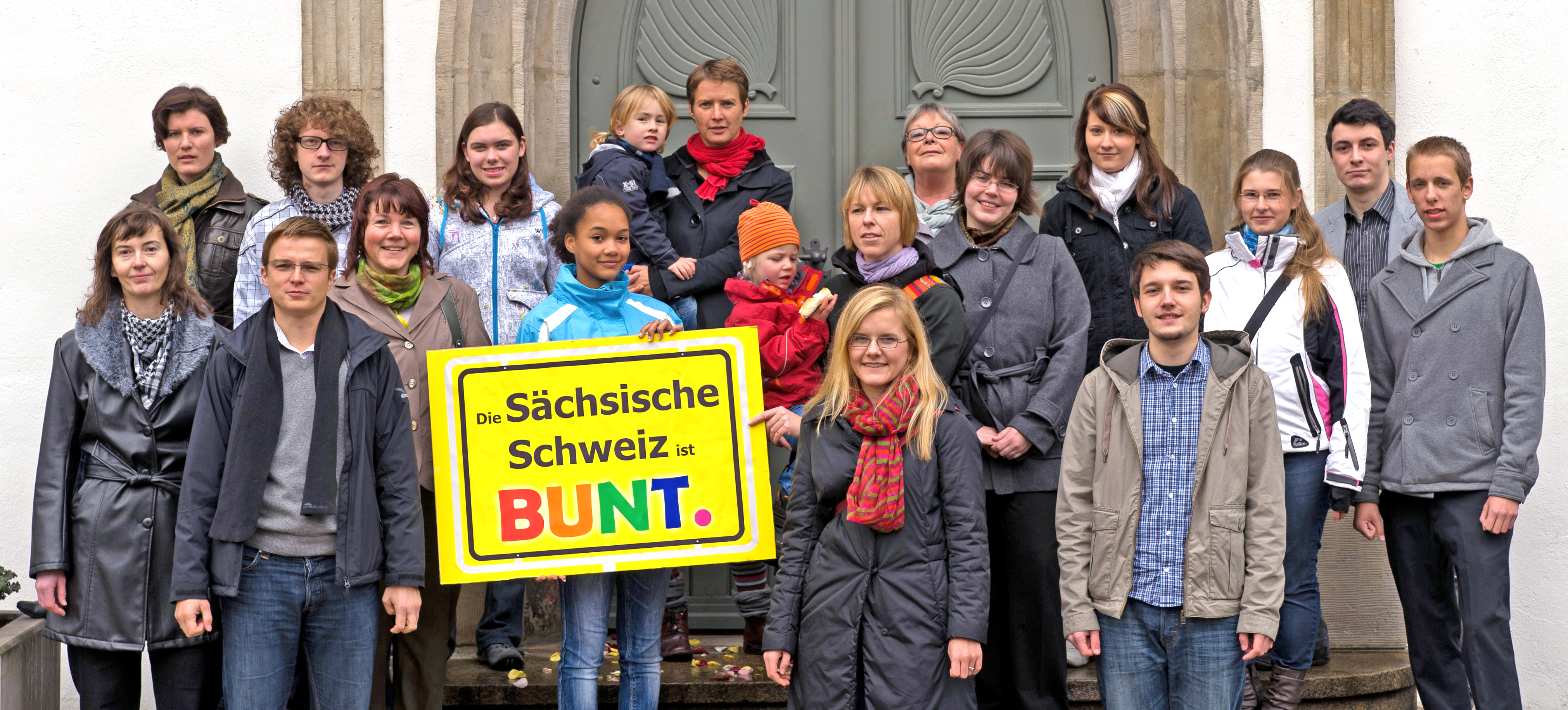 KI generiert: Das Bild zeigt eine Gruppe von Menschen, die vor einem Gebäude stehen und gemeinsam ein Schild mit der Aufschrift "Die Sächsische Schweiz ist BUNT." halten. Der Hauptinhalt des Bildes ist die Darstellung von Vielfalt und Zusammenhalt in der Sächsischen Schweiz.