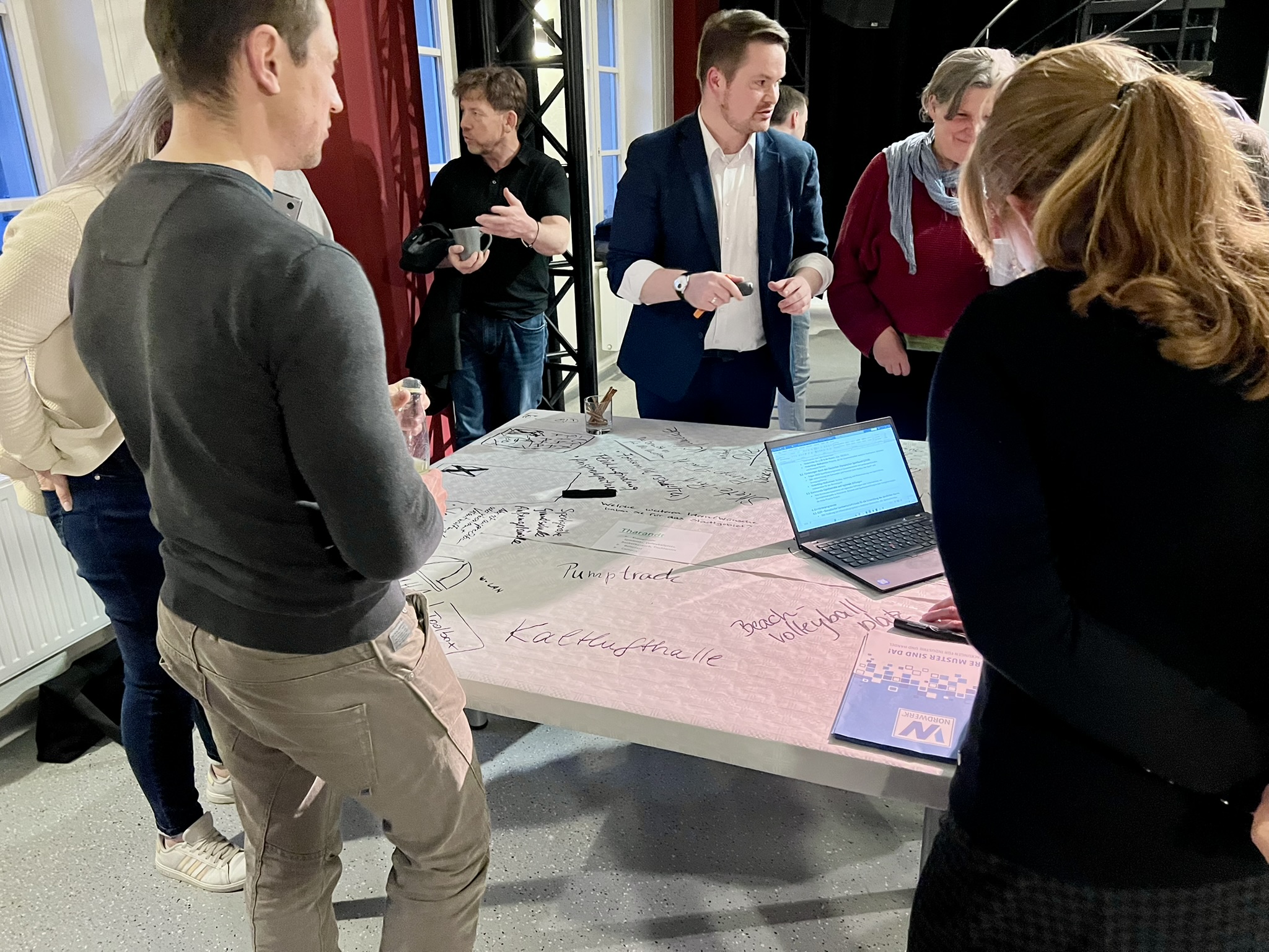 KI generiert: Das Bild zeigt eine Gruppe von Personen, die sich um einen großen Tisch versammelt haben, auf dem ein Plan oder eine Karte ausgebreitet ist. Der Hauptinhalt des Bildes ist eine Besprechung oder ein Workshop, bei dem Ideen oder Strategien diskutiert werden.