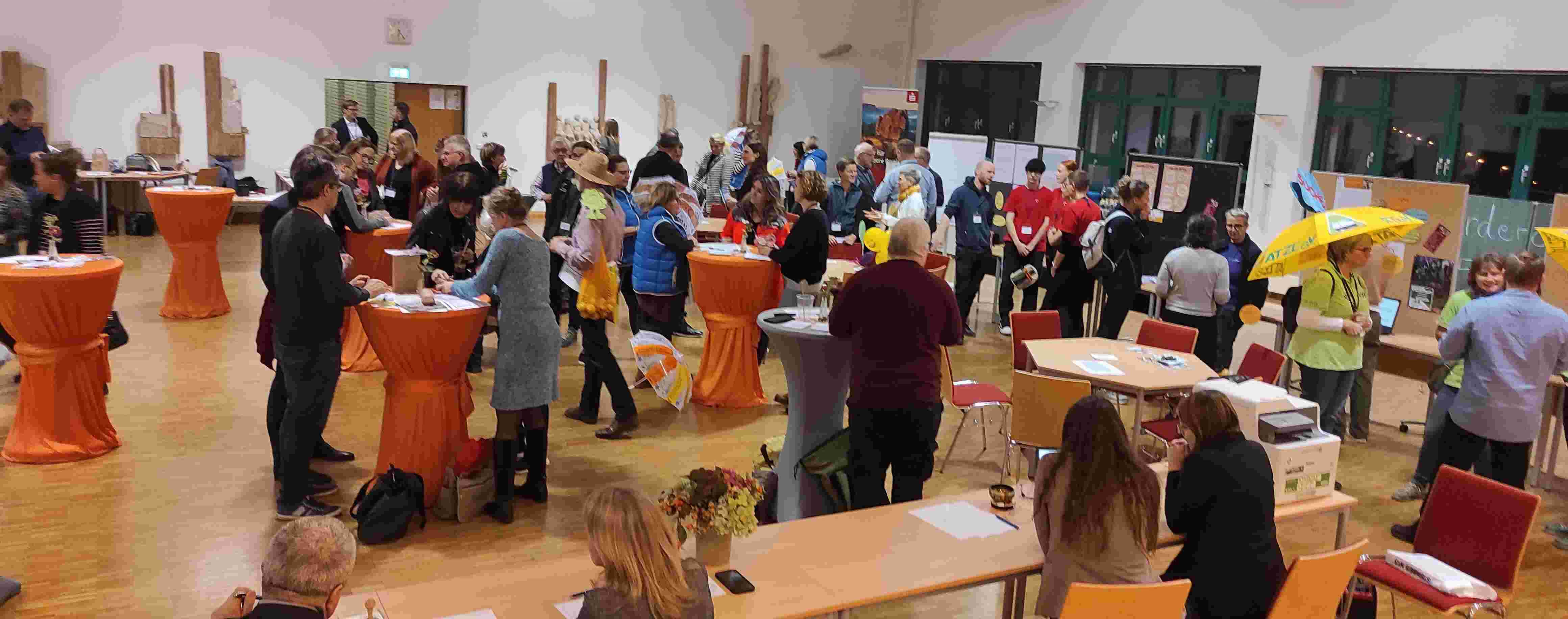 KI generiert: Das Bild zeigt eine belebte Indoor-Veranstaltung mit vielen Menschen, die an Stehtischen mit orangefarbenen Tischdecken stehen und sich unterhalten. Im Vordergrund sind einige Personen an Tischen sitzend zu sehen, während im Hintergrund weitere Besucher und Informationsstände erkennbar sind. Der Hauptinhalt des Bildes ist eine gesellige Versammlung oder Messe.