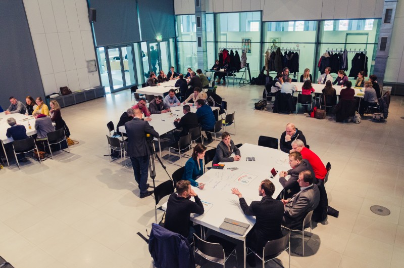 KI generiert: Das Bild zeigt eine Gruppe von Personen, die in kleinen Gruppen an mehreren Tischen in einem großen, modernen Raum mit hohen Fenstern und hellem Licht zusammenarbeiten oder diskutieren. Der Hauptinhalt des Bildes ist eine Arbeits- oder Meeting-Situation, bei der die Teilnehmer aktiv in Gespräche oder Aktivitäten eingebunden sind.