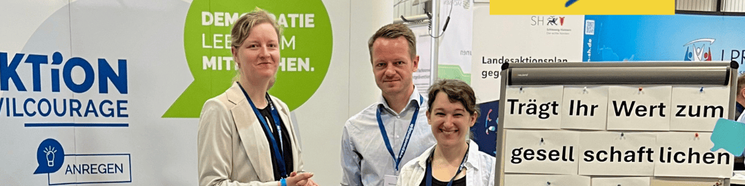 KI generiert: Das Bild zeigt drei Personen, die vor einem Stand mit dem Schriftzug "Aktion Zivilcourage" und einem Slogan zur Demokratie stehen. Im Hintergrund sind Informationsmaterialien und ein Bildschirm mit dem Text "Trägt Ihr Wert zum gesellschaftlichen Leben bei?" zu sehen.