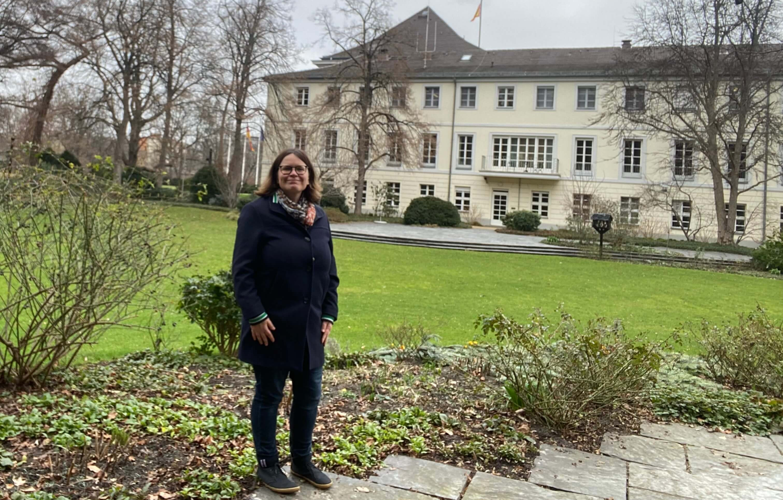 KI generiert: Das Bild zeigt eine Frau, die vor einem großen, mehrstöckigen Gebäude mit vielen Fenstern steht, umgeben von einem gepflegten Garten mit Rasen, Sträuchern und Bäumen. Die Frau trägt eine dunkle Jacke, eine Brille und ein Schal und steht auf einem Steinweg.