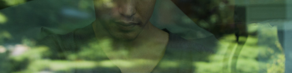 KI generiert: Das Bild zeigt eine Person mit einem neutralen Gesichtsausdruck, die durch eine Glasfläche oder ein Fenster zu sehen ist. Im Hintergrund sind verschwommene grüne Bäume und eine unscharfe Landschaft zu erkennen.