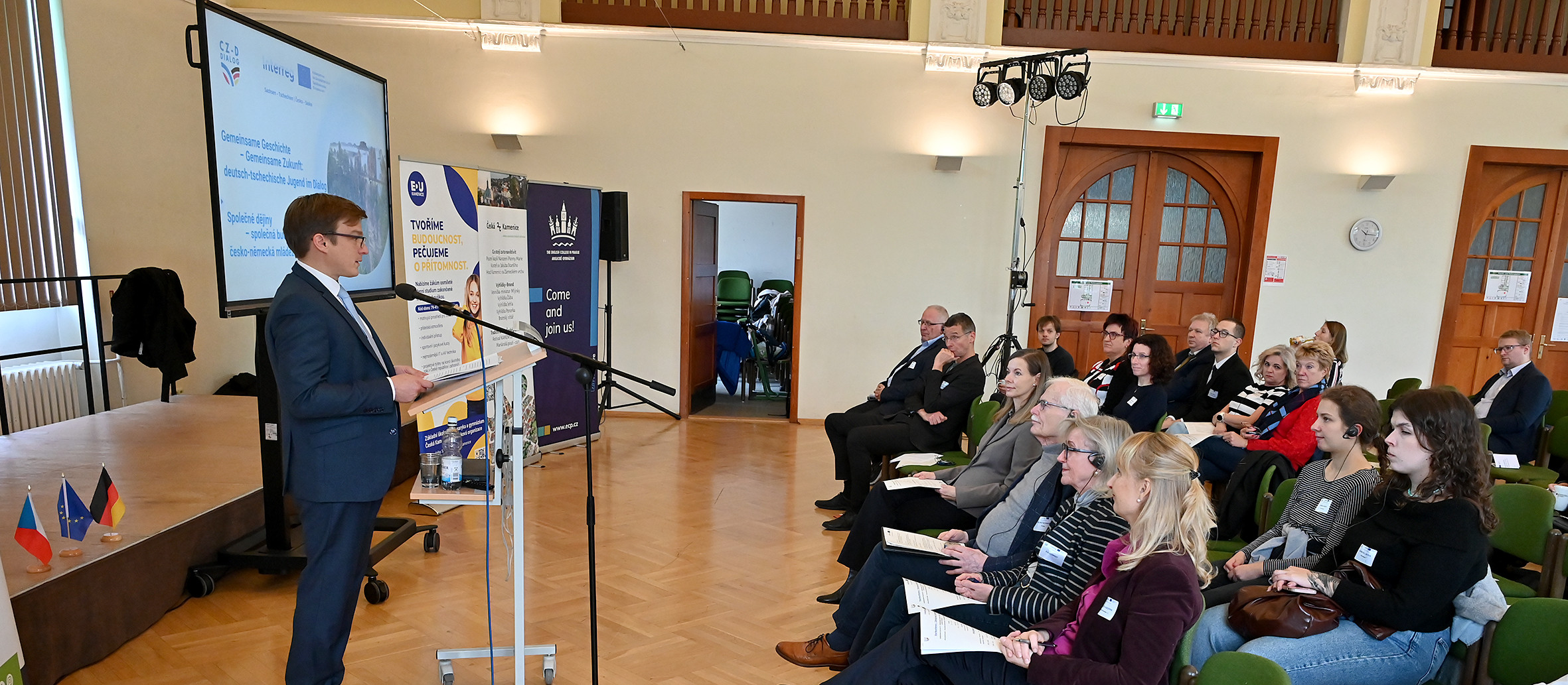 KI generiert: Das Bild zeigt eine Präsentation in einem Konferenzraum, bei der ein Mann vor einem Publikum spricht. Der Hauptinhalt ist eine formelle Veranstaltung oder Konferenz mit Fokus auf den Vortragenden und die Zuhörer.
