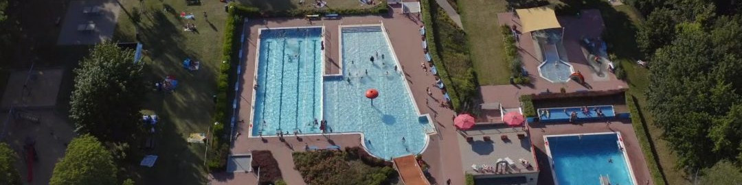 KI generiert: Das Bild zeigt eine Luftaufnahme eines Schwimmbads mit mehreren Becken, umgeben von Grünflächen und Gebäuden. Der Hauptinhalt des Bildes ist das große, belebte Freibad mit verschiedenen Wasserbecken und Liegeflächen.