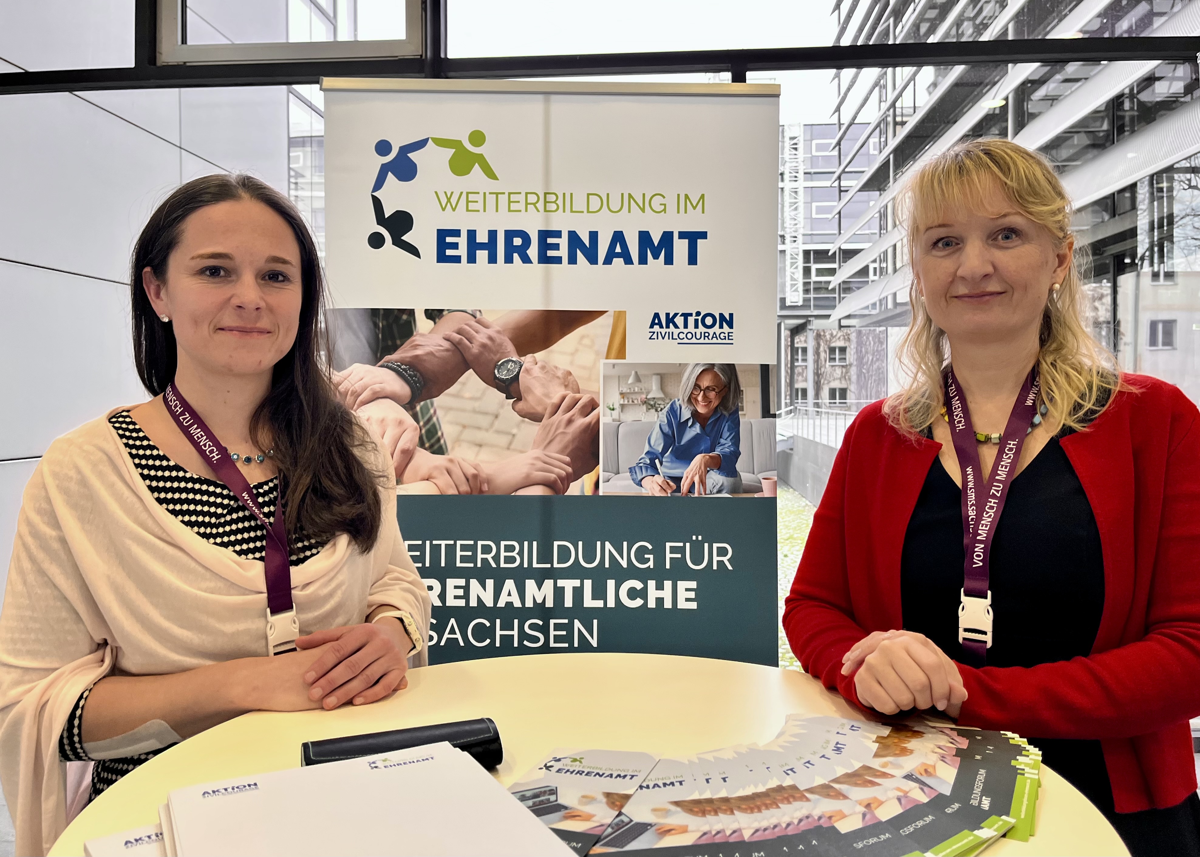 KI generiert: Das Bild zeigt zwei Frauen an einem Tisch mit Informationsmaterial und einem Banner, das für Weiterbildung im Ehrenamt wirbt. Der Hauptinhalt des Bildes ist die Bewerbung von Weiterbildungsmöglichkeiten für ehrenamtliche Tätigkeiten.