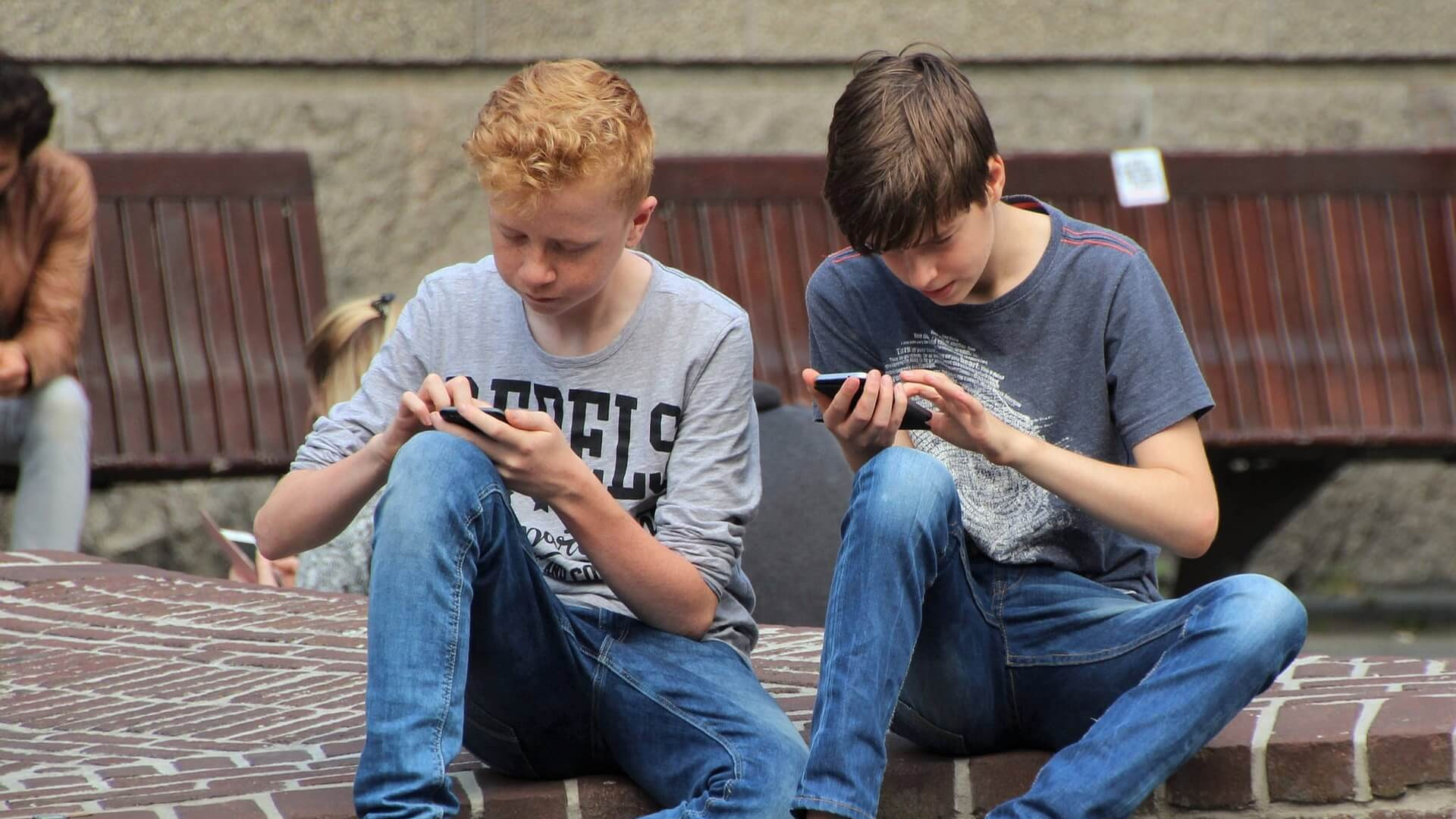 KI generiert: Das Bild zeigt zwei Jugendliche, die auf einer Bank sitzen und jeweils auf ihr Smartphone schauen. Der Hauptinhalt des Bildes ist die Nutzung von Smartphones durch die Jugendlichen in einer öffentlichen Umgebung.
