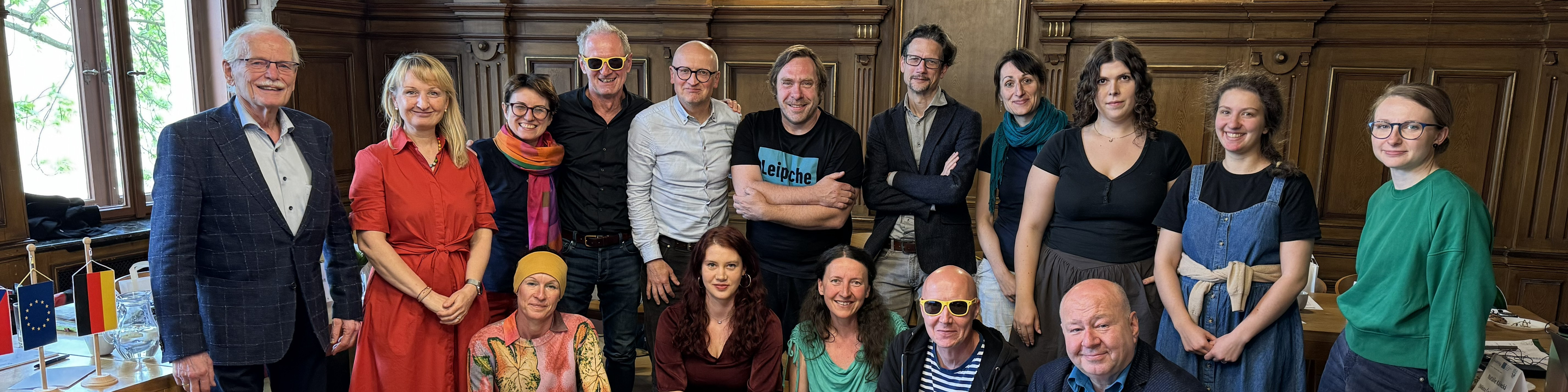 KI generiert: Das Bild zeigt eine Gruppe von Personen, die in einem Raum mit Holzvertäfelung und großen Fenstern posieren. Im Vordergrund sind zwei kleine Flaggen zu sehen, was darauf hindeuten könnte, dass es sich um eine offizielle oder internationale Zusammenkunft handelt.