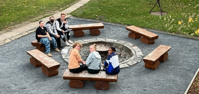KI generiert: Das Bild zeigt eine Gruppe von sechs Personen, die um ein zentrales Feuer in einer Feuerstelle im Freien sitzen. Sie sind auf rustikalen Holzbänken platziert, die in einem Kreis um die Feuerstelle angeordnet sind.