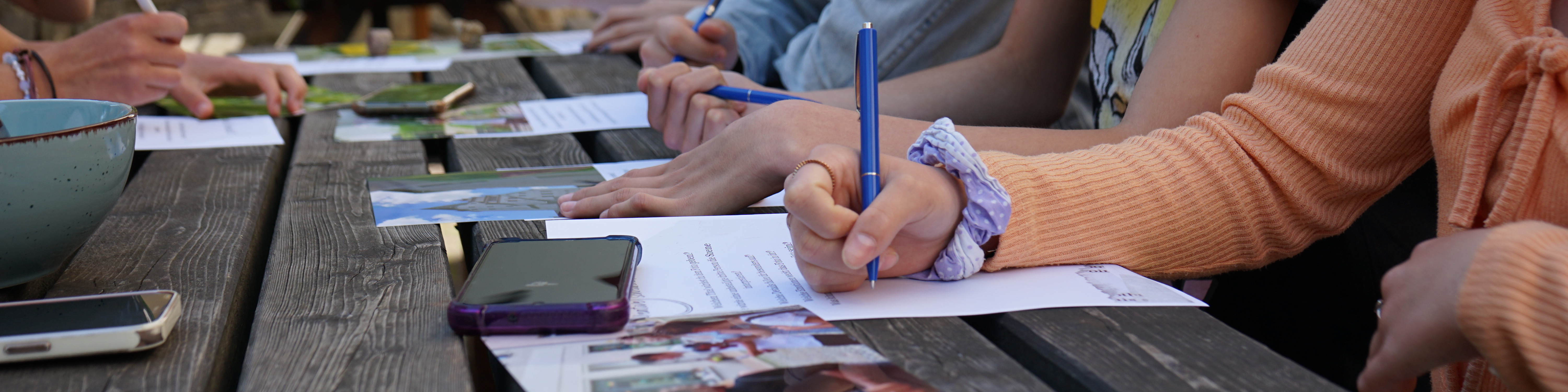 KI generiert: Das Bild zeigt eine Gruppe von Personen, die an einem Holztisch sitzen und auf Papier schreiben oder zeichnen. Der Hauptinhalt des Bildes ist eine kreative oder pädagogische Aktivität, bei der die Teilnehmer mit Stiften und Smartphones arbeiten.