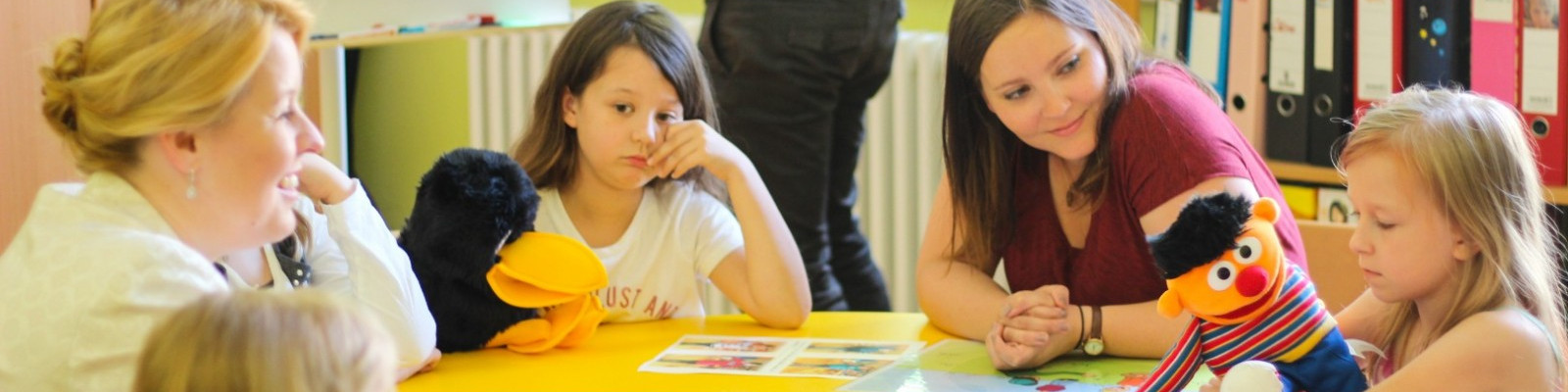 KI generiert: Das Bild zeigt eine Gruppe von Kindern und Erwachsenen, die an einem Tisch sitzen und mit Spielzeug und Bildern spielen oder arbeiten. Der Hauptinhalt des Bildes ist eine interaktive und pädagogische Aktivität, möglicherweise in einem Kindergarten oder einer Vorschule.