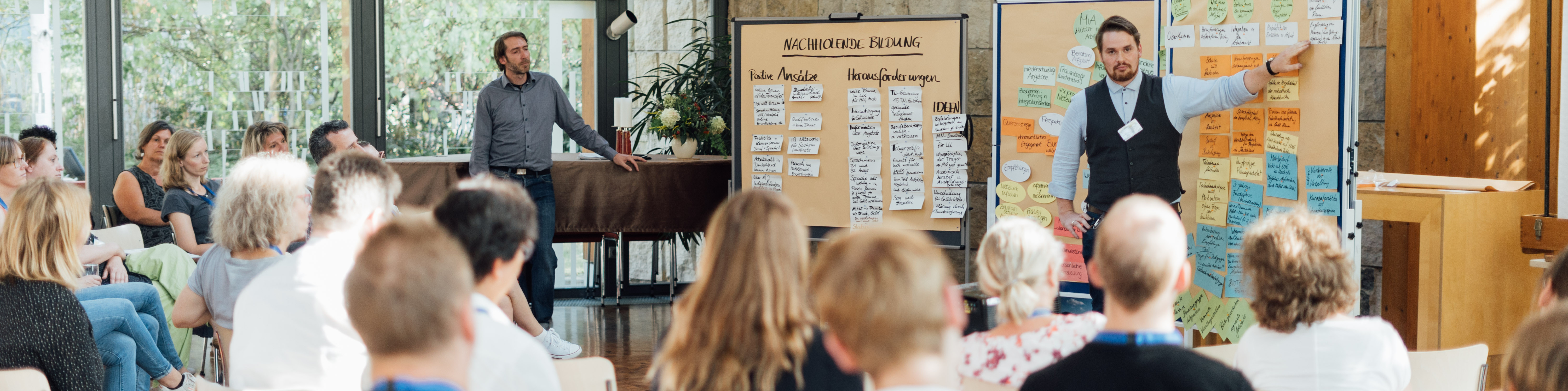 KI generiert: Das Bild zeigt eine Gruppe von Menschen in einem Seminarraum, wobei zwei Männer vorne stehen und Präsentationen halten. Der Hauptinhalt des Bildes ist eine interaktive Schulung oder ein Workshop, bei dem Ideen und Informationen auf Flipcharts präsentiert und diskutiert werden.