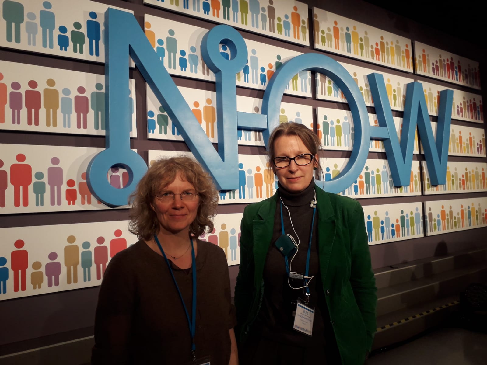 KI generiert: Das Bild zeigt zwei Frauen, die vor einer Wand mit großen, blauen Buchstaben stehen, die das Wort "NOW" bilden. Im Hintergrund sind bunte Piktogramme von Menschen zu sehen.