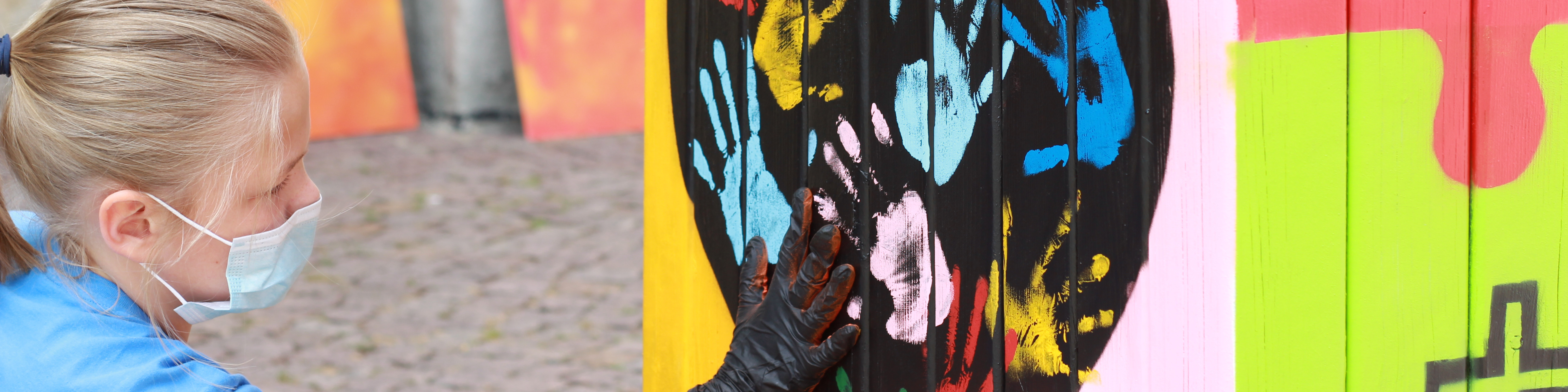 KI generiert: Das Bild zeigt eine Person mit einer Maske und Handschuhen, die eine bunte Graffiti-Wand mit Handabdrücken berührt. Der Hauptinhalt des Bildes ist die Interaktion mit der farbenfrohen Street-Art.