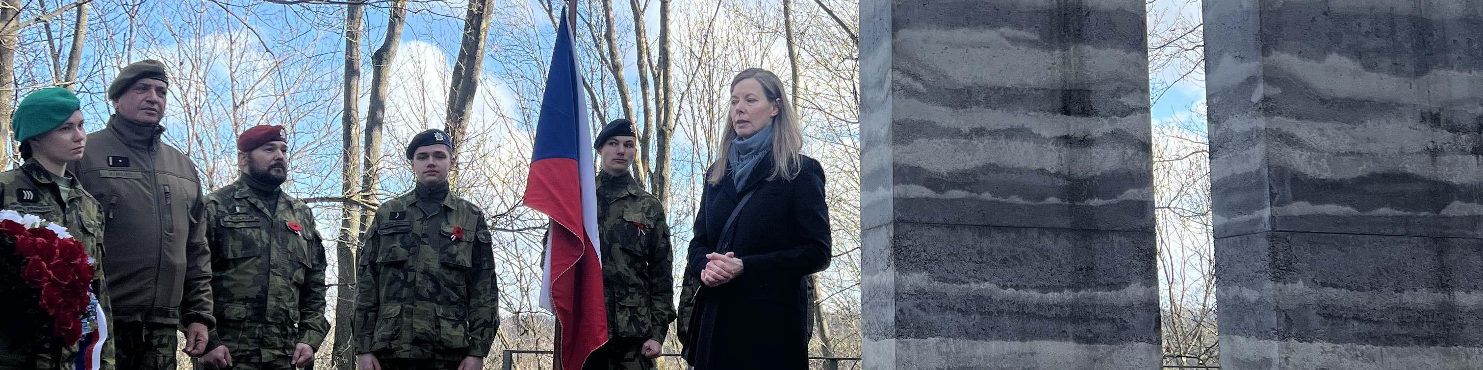 KI generiert: Das Bild zeigt eine Gruppe von Soldaten in Uniform, die an einer Gedenkveranstaltung teilnehmen, wobei eine Frau eine Rede vor einem Denkmal hält. Im Hintergrund ist eine Flagge zu sehen, die zwischen den Bäumen weht.