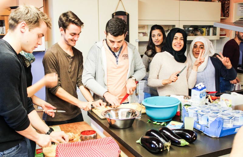 KI generiert: Das Bild zeigt eine Gruppe von Menschen, die gemeinsam in einer Küche kochen und sich dabei unterhalten. Im Vordergrund sind verschiedene Zutaten wie Auberginen und Joghurtbecher zu sehen.