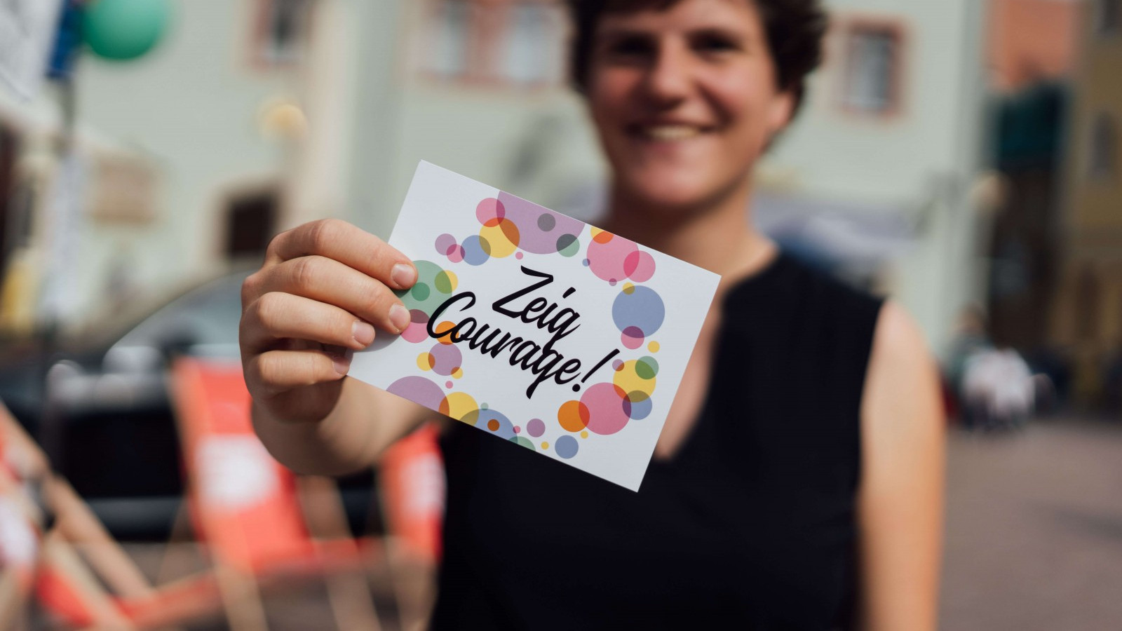 KI generiert: Das Bild zeigt eine lächelnde Person, die eine Karte mit der Aufschrift "Zeig Courage!" und bunten Kreisen in der Hand hält. Im Hintergrund sind unscharf ein Straßenfest oder eine öffentliche Veranstaltung mit Stühlen und Gebäuden zu erkennen.
