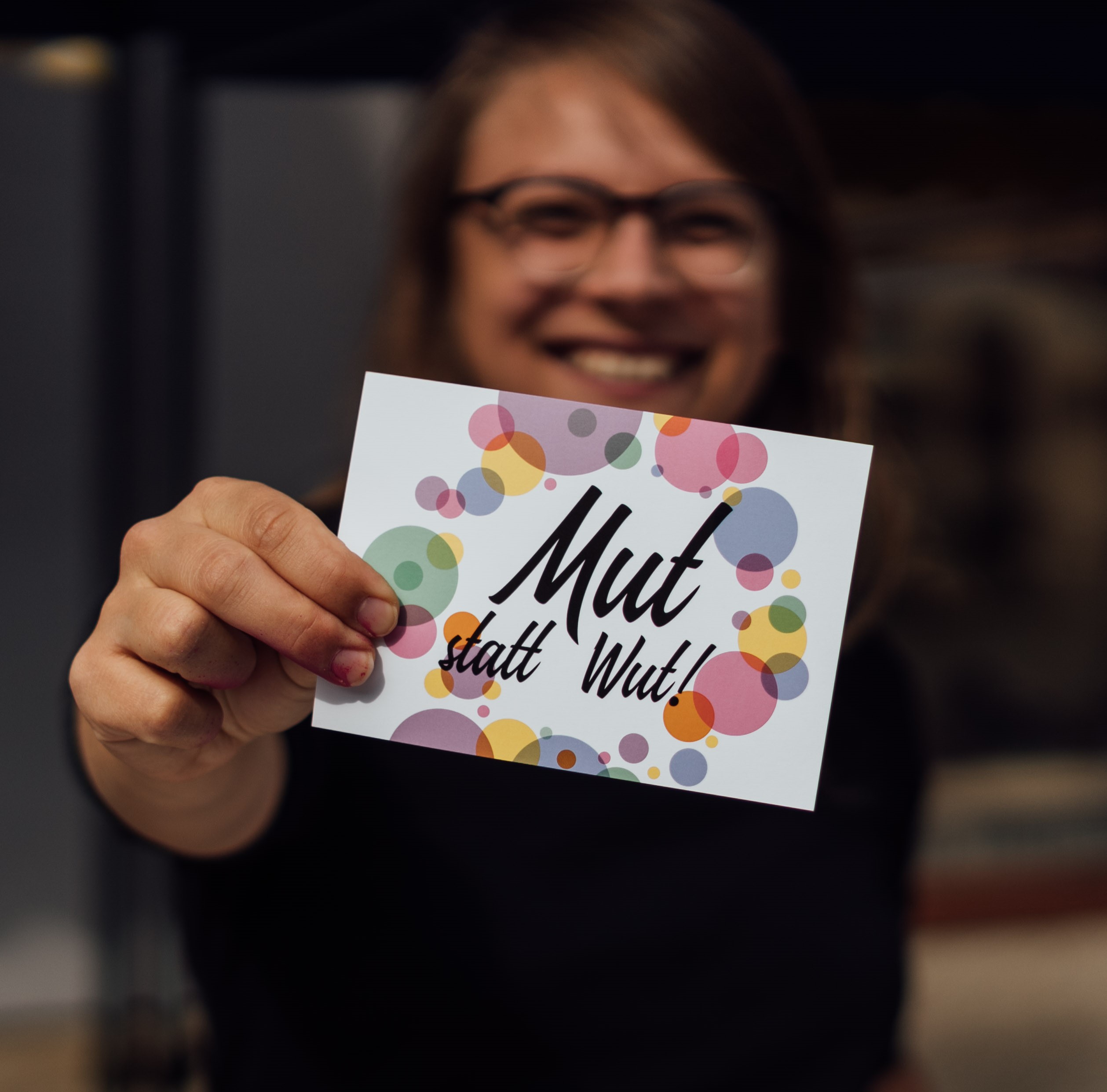 KI generiert: Das Bild zeigt eine Person, die ein weißes Kartenblatt mit bunten Kreisen und der Aufschrift "Mut statt Wut!" in der Hand hält. Der Hauptinhalt des Bildes ist die Botschaft auf der Karte, die Mut und Positivität fördert.