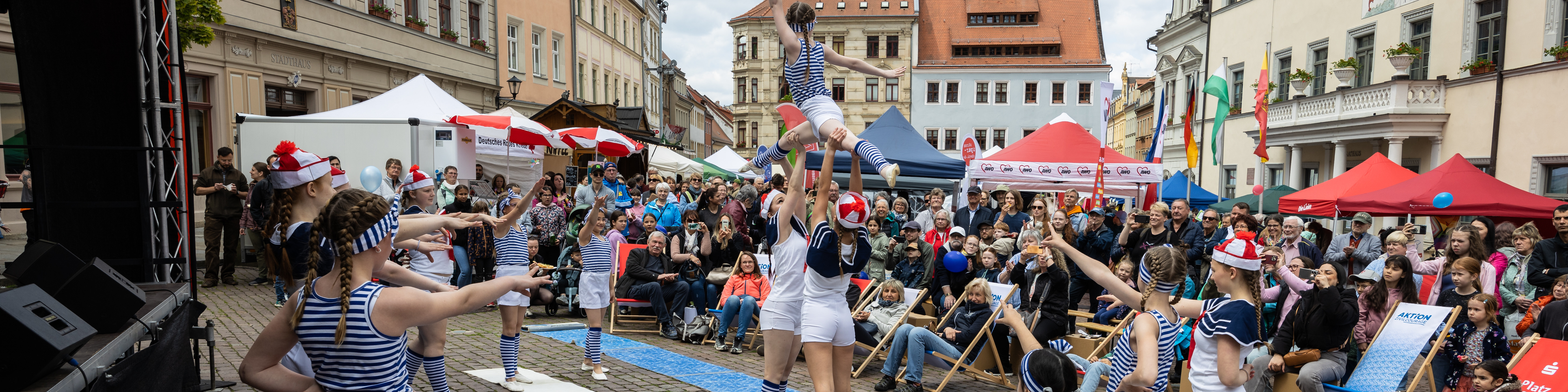 KI generiert: Das Bild zeigt eine belebte Straßenveranstaltung mit Akrobaten, die eine menschliche Pyramide bilden, während ein Publikum zuschaut. Im Hintergrund sind bunte Zelte und historische Gebäude zu sehen.