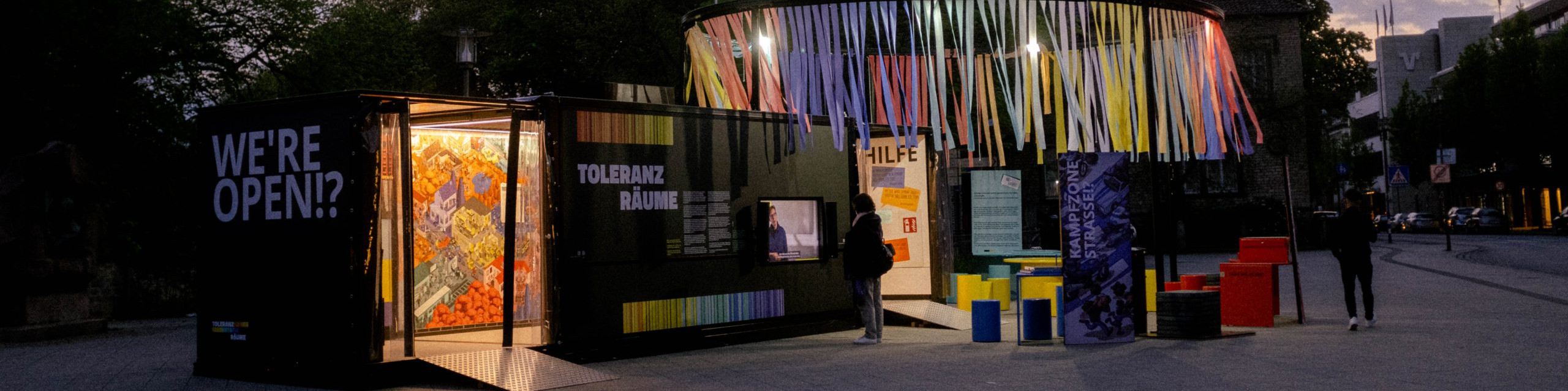 KI generiert: Das Bild zeigt eine moderne, farbenfrohe Installation oder Ausstellung im Freien mit dem Schriftzug "WE'RE OPEN!" und "TOLERANZ RÄUME". Der Hauptinhalt ist eine kreative, interaktive Kunstinstallation, die Passanten einlädt, sie zu erkunden.