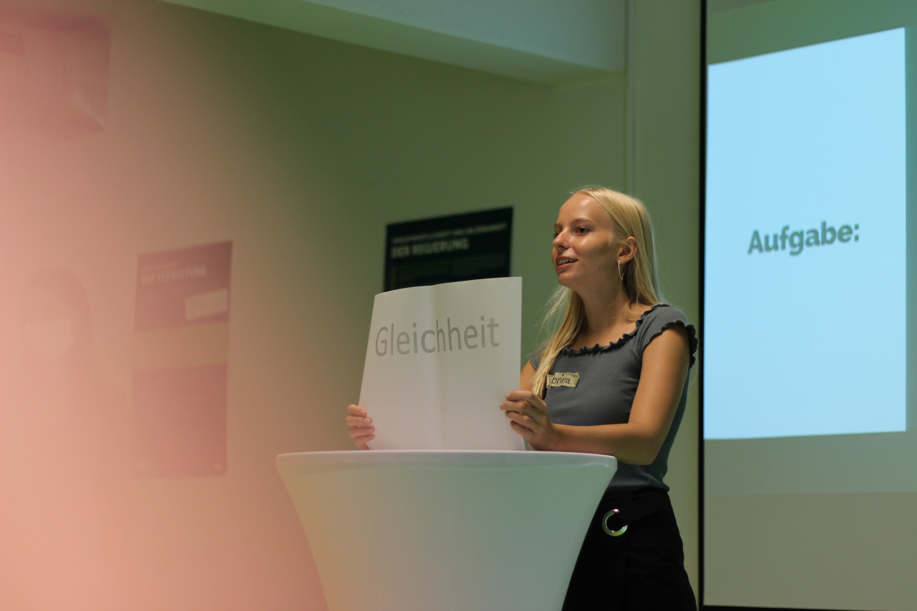 KI generiert: Das Bild zeigt eine Frau, die an einem Pult steht und ein Schild mit der Aufschrift "Gleichheit" hochhält. Im Hintergrund ist eine Leinwand mit dem Wort "Aufgabe:" zu sehen.
