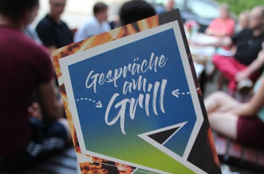 KI generiert: Das Bild zeigt eine Gruppe von Menschen, die sich im Freien versammelt haben, mit einem Schild im Vordergrund, auf dem "Gespräche am Grill" steht. Der Hauptinhalt des Bildes ist die Ankündigung oder der Titel einer Veranstaltung, die wahrscheinlich eine gesellige Zusammenkunft mit Gesprächen beim Grillen ist.
