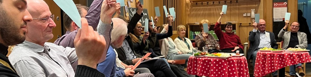 KI generiert: Das Bild zeigt eine Gruppe von Personen in einem Raum, die ihre Hände mit blauen Karten hochhalten. Es scheint sich um eine Abstimmung oder eine Wahl zu handeln.