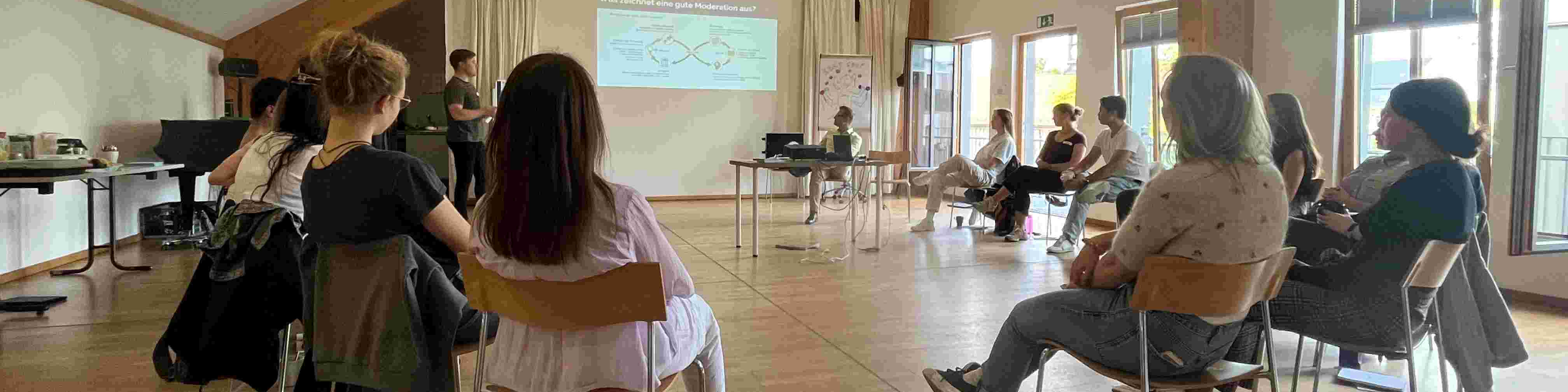 KI generiert: Das Bild zeigt eine Gruppe von Personen in einem Seminarraum, wobei eine Person vorne steht und eine Präsentation hält. Der Hauptinhalt des Bildes ist eine Bildungs- oder Schulungsveranstaltung.