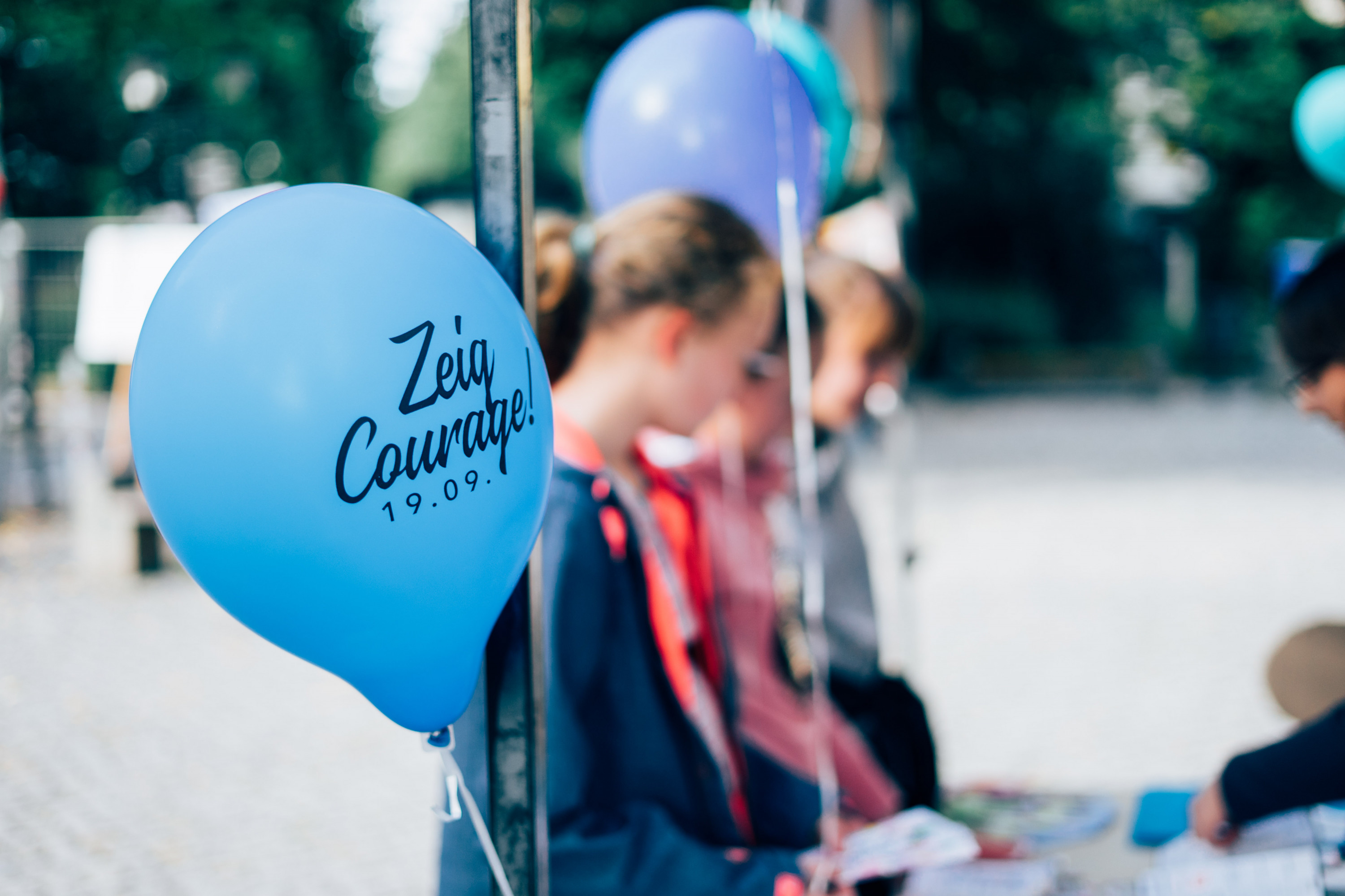 KI generiert: Das Bild zeigt einen blauen Luftballon mit der Aufschrift "Zeig Courage! 19.09." im Vordergrund, während im Hintergrund unscharf einige Personen zu sehen sind, die möglicherweise an einer Veranstaltung teilnehmen. Der Hauptinhalt des Bildes ist der Luftballon mit der Botschaft, die auf eine mutige Handlung oder ein Event hinweist.