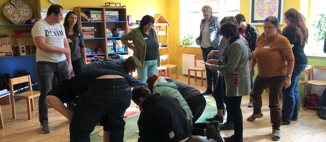 KI generiert: Das Bild zeigt eine Gruppe von Erwachsenen in einem Raum, der wie ein Klassenzimmer oder ein Spielzimmer für Kinder eingerichtet ist. Einige Personen beugen sich über eine Person, die auf dem Boden liegt, möglicherweise im Rahmen einer Übung oder eines Spiels.