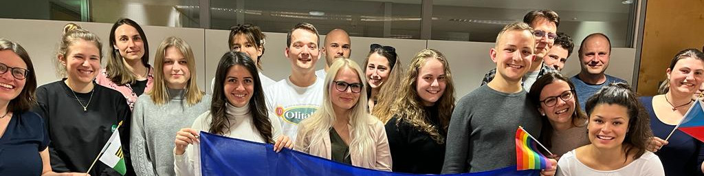 KI generiert: Das Bild zeigt eine Gruppe von Menschen, die gemeinsam posieren und dabei verschiedene Flaggen halten, darunter eine Regenbogenflagge und eine blau-weiße Flagge. Der Hauptinhalt des Bildes ist die Darstellung einer vielfältigen und fröhlichen Gruppe, die möglicherweise an einem Event oder einer Feier teilnimmt.