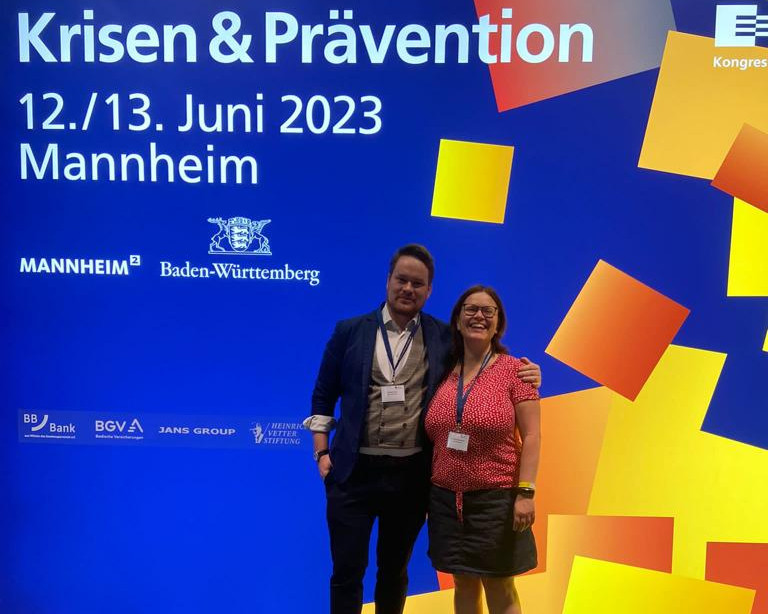 KI generiert: Das Bild zeigt eine Gruppe von Personen auf einer Messe oder Ausstellung, die sich an einem Stand unterhalten. Der Stand hat mehrere Poster mit Informationen, darunter Themen wie Verkehrssicherheit und Präventionsrat im Freistaat Sachsen.