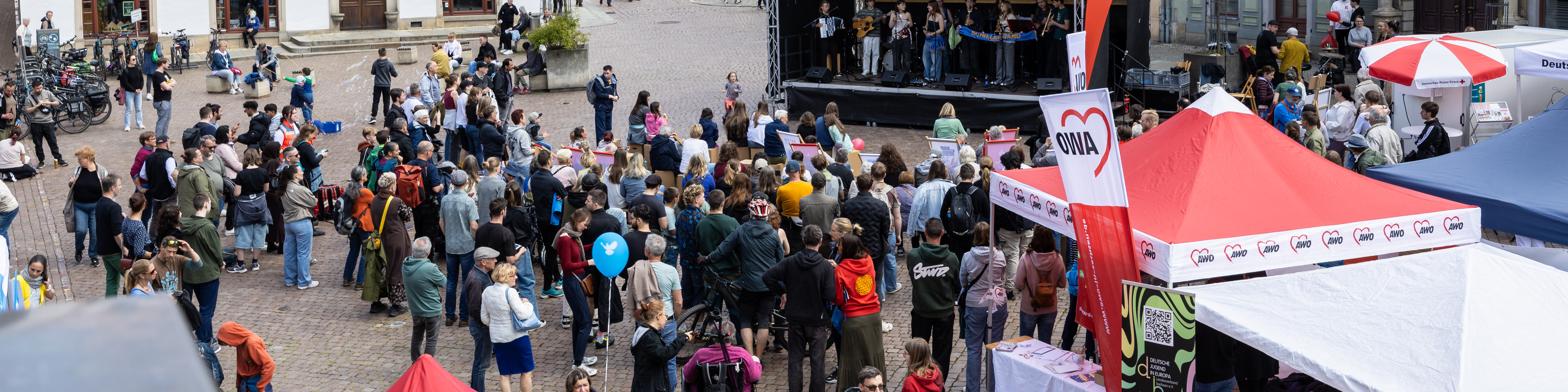 KI generiert: Das Bild zeigt eine belebte öffentliche Veranstaltung mit vielen Menschen, die sich um eine Bühne und verschiedene Stände versammeln. Im Vordergrund sind Zelte mit Aufschriften wie "OWA" und "Caritas" zu sehen, was auf eine gemeinnützige oder soziale Veranstaltung hindeuten könnte.