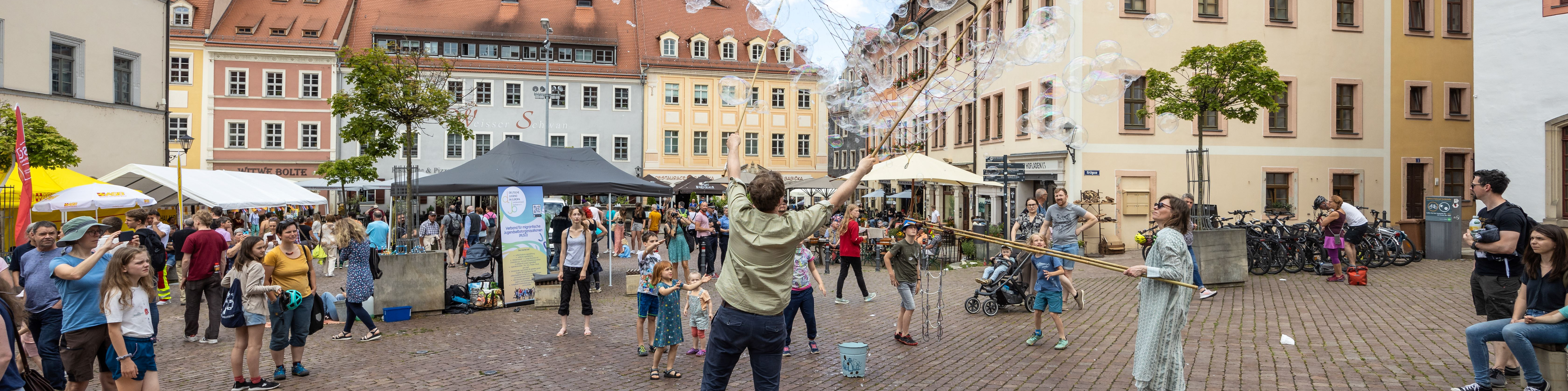 KI generiert: Das Bild zeigt eine belebte Platzszene mit Menschen, die sich an verschiedenen Aktivitäten beteiligen, darunter das Erzeugen großer Seifenblasen. Im Hintergrund sind bunte Gebäude und Marktstände zu sehen, die eine lebendige Atmosphäre schaffen.