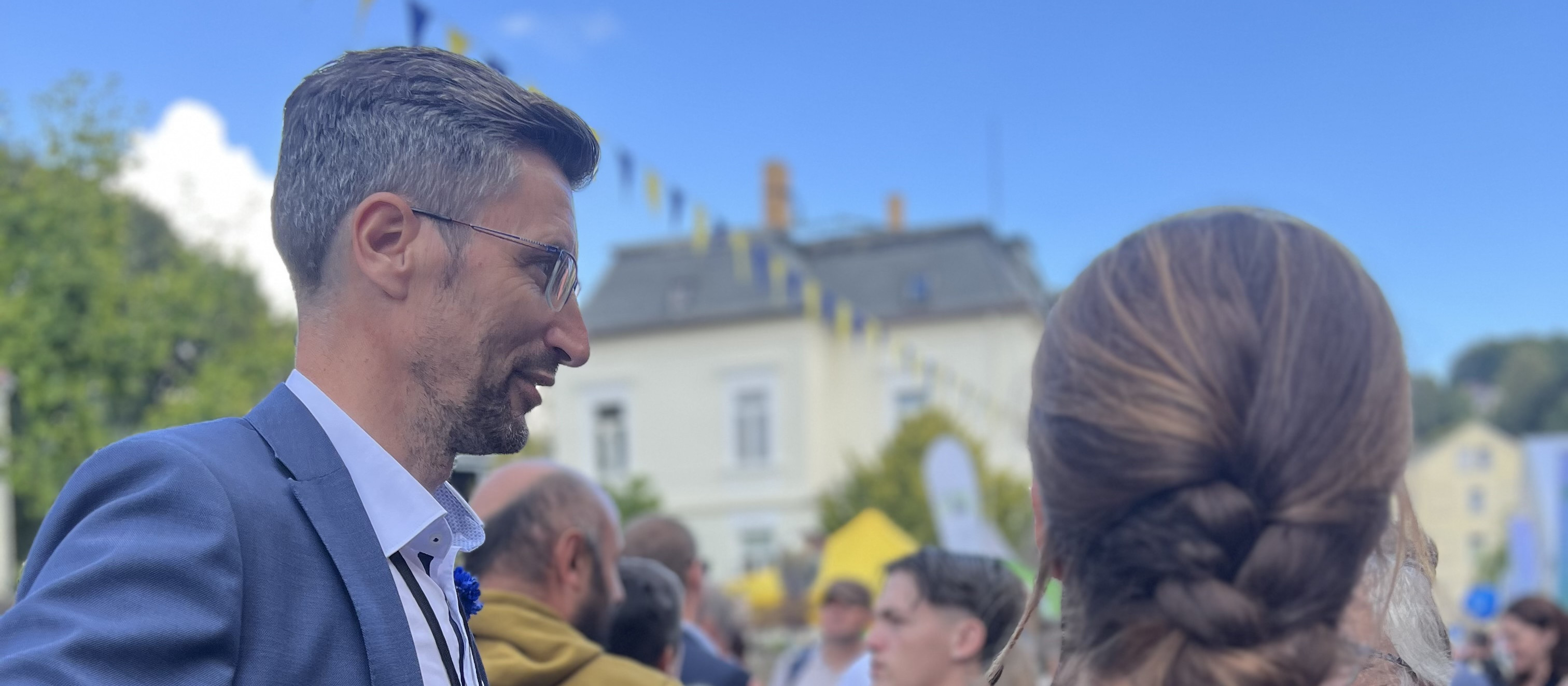 KI generiert: Das Bild zeigt eine Gruppe von Menschen, die sich im Freien unterhalten, mit einem Mann im Vordergrund, der eine Brille und ein Sakko trägt. Im Hintergrund sind Gebäude und ein klarer blauer Himmel zu sehen.