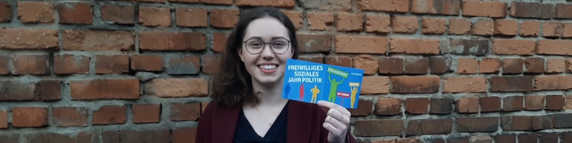KI generiert: Das Bild zeigt eine lächelnde Person mit Brille, die vor einer Ziegelsteinmauer steht und ein Buch mit dem Titel "Freiwilliges Soziales Jahr Politik" in der Hand hält. Der Hauptinhalt des Bildes ist die Präsentation des Buches.